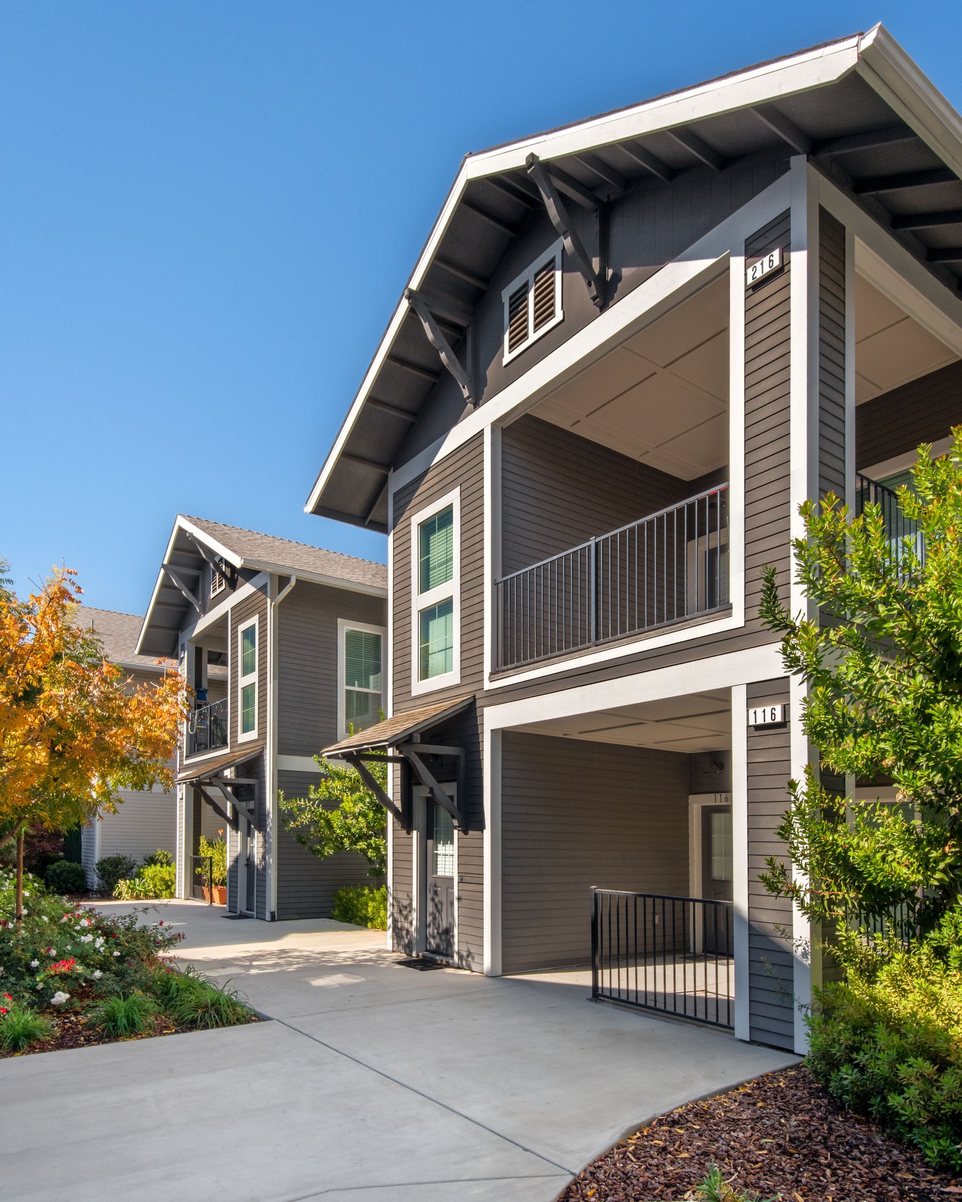 Two-story gray apartment buildings with balconies, blue sky, and greenery along a concrete path.