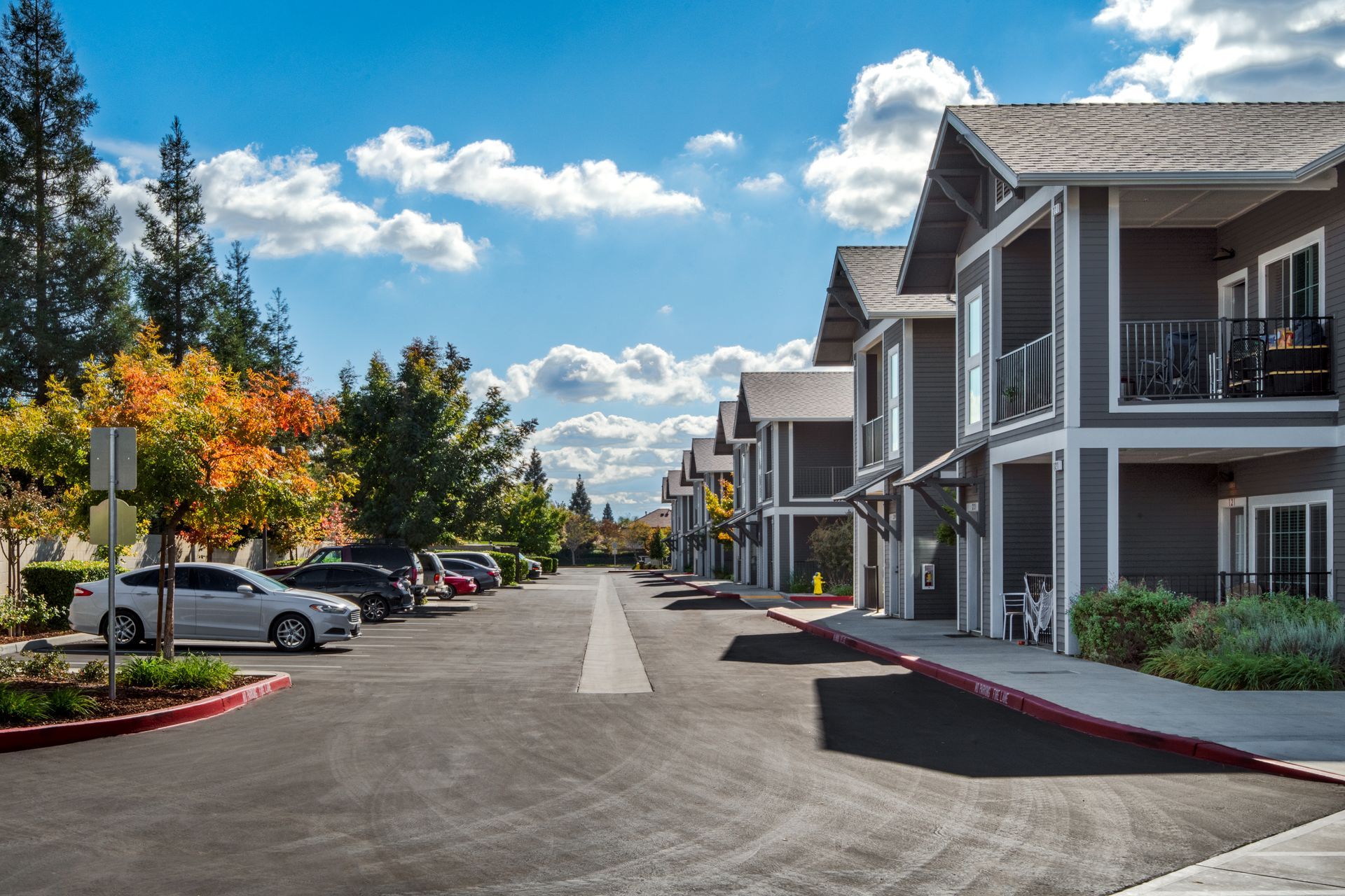 Apartment complex with parking area under a blue sky with puffy clouds; autumn leaves and cars.