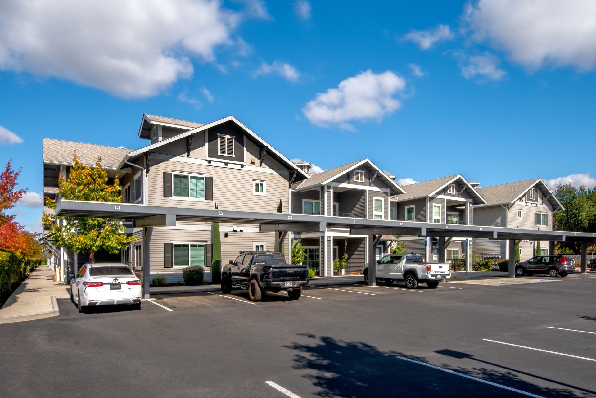 Apartment complex with covered parking, light grey siding, and blue sky with puffy clouds.