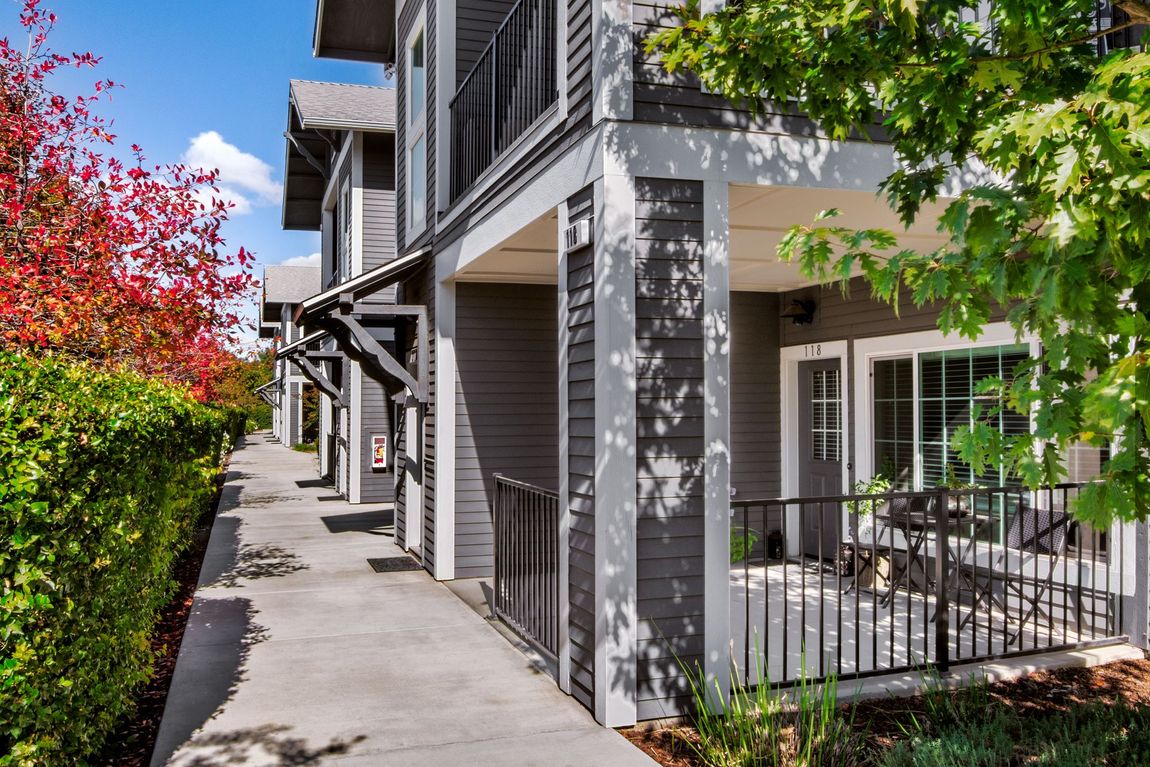 Gray townhomes with balconies, sidewalk, and greenery. Sunny day.