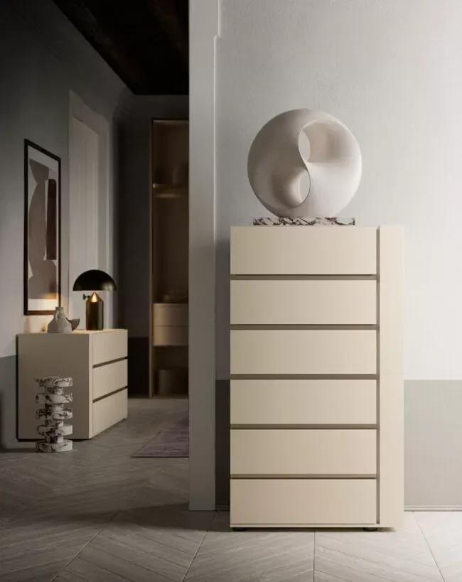 Comò color crema con sei cassetti, sormontato da una scultura sferica. Un altro comò sullo sfondo, vicino a una lampada e a un'opera d'arte.