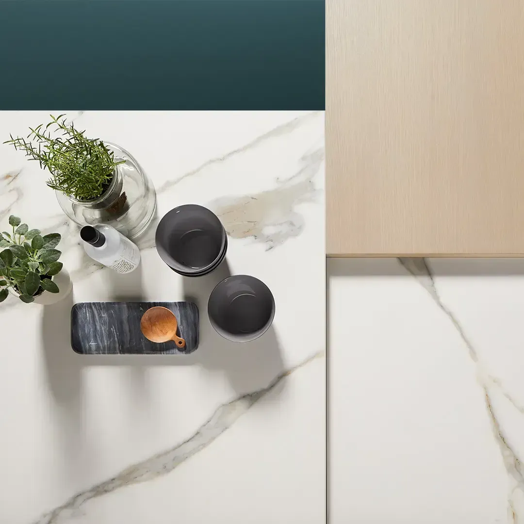 Ripresa dall'alto di un tavolo con motivi in marmo, su cui sono disposti utensili da cucina come piante, ciotole e un mortaio con pestello. Sopra, una superficie color verde acqua e legno.