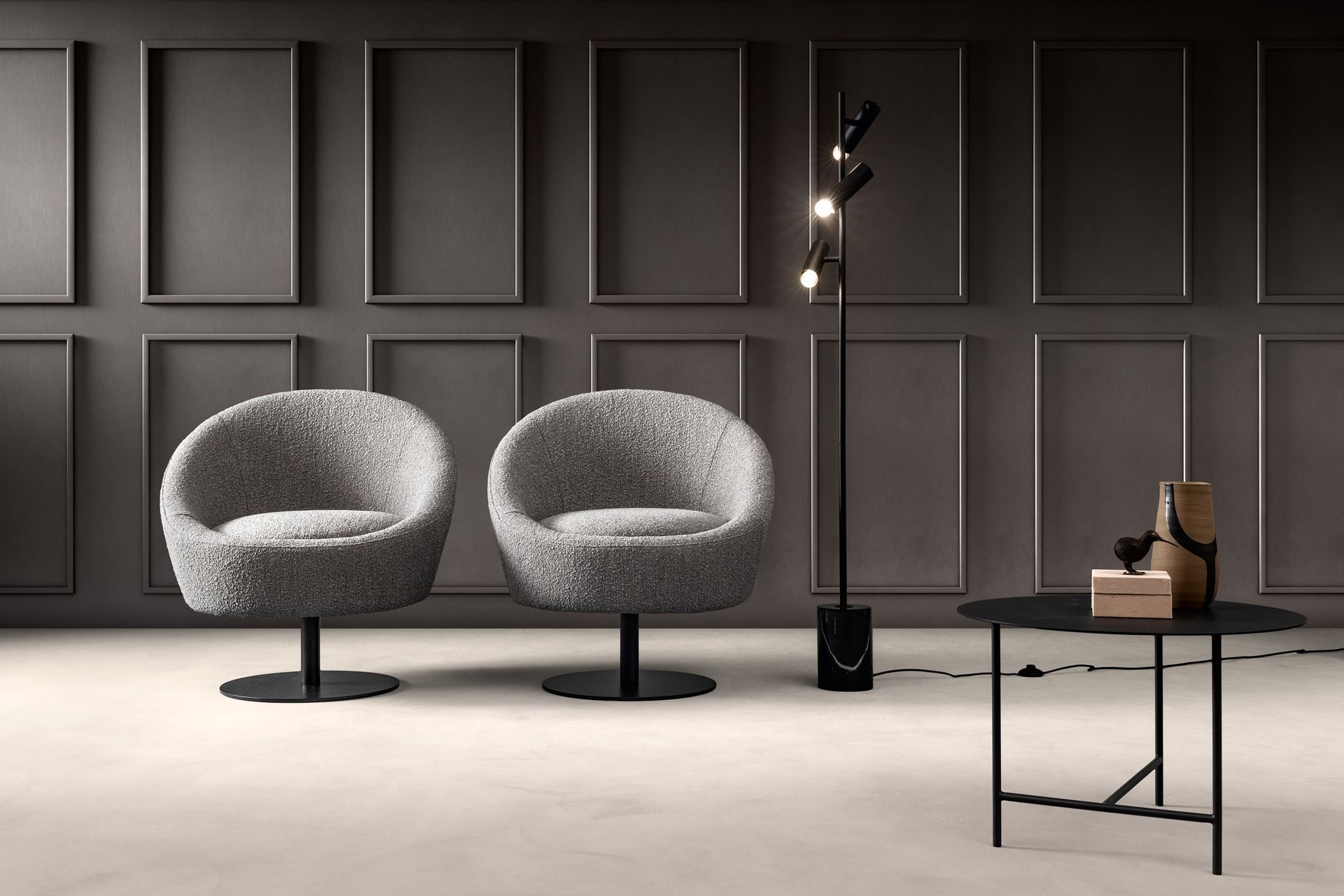 Due sedie grigie pelose, una lampada da terra nera e un tavolino laterale contro una parete grigio scuro con pannelli rettangolari.