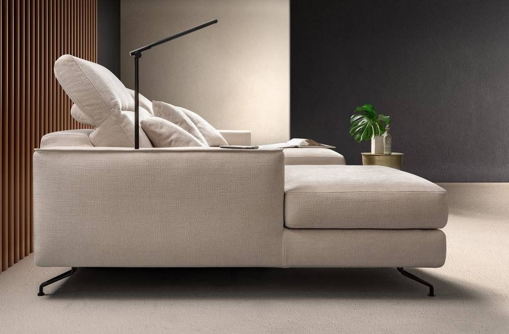 Divano componibile moderno beige con poggiatesta regolabili e lampada nera integrata, contro una parete neutra con una pianta.
