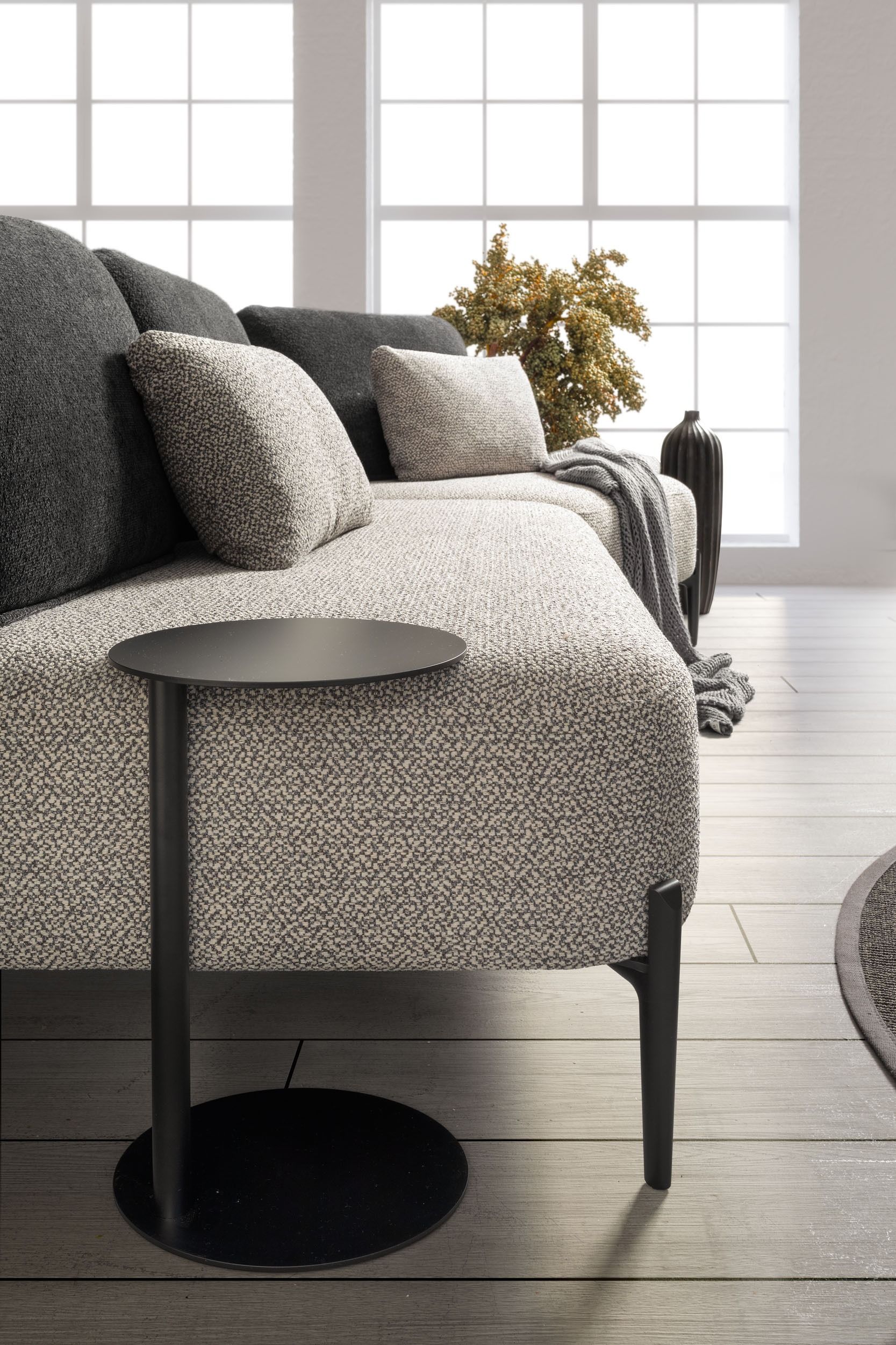 Un divano con tessuto grigio testurizzato e cuscini scuri accanto a un tavolino nero. Dietro c'è una finestra illuminata.