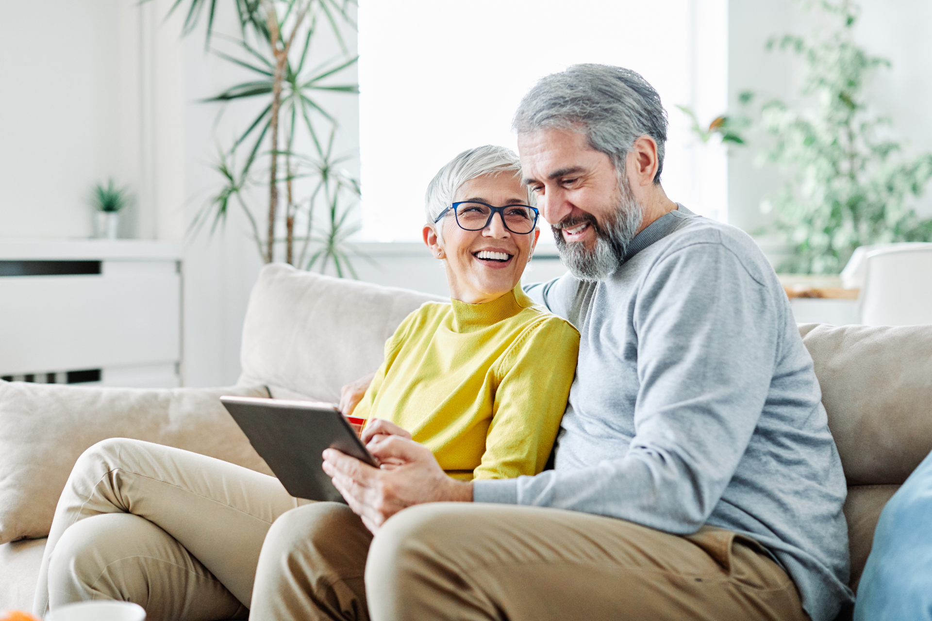 Couple on a sofa looking at a tablet, smiling. They are in a well-lit living room.