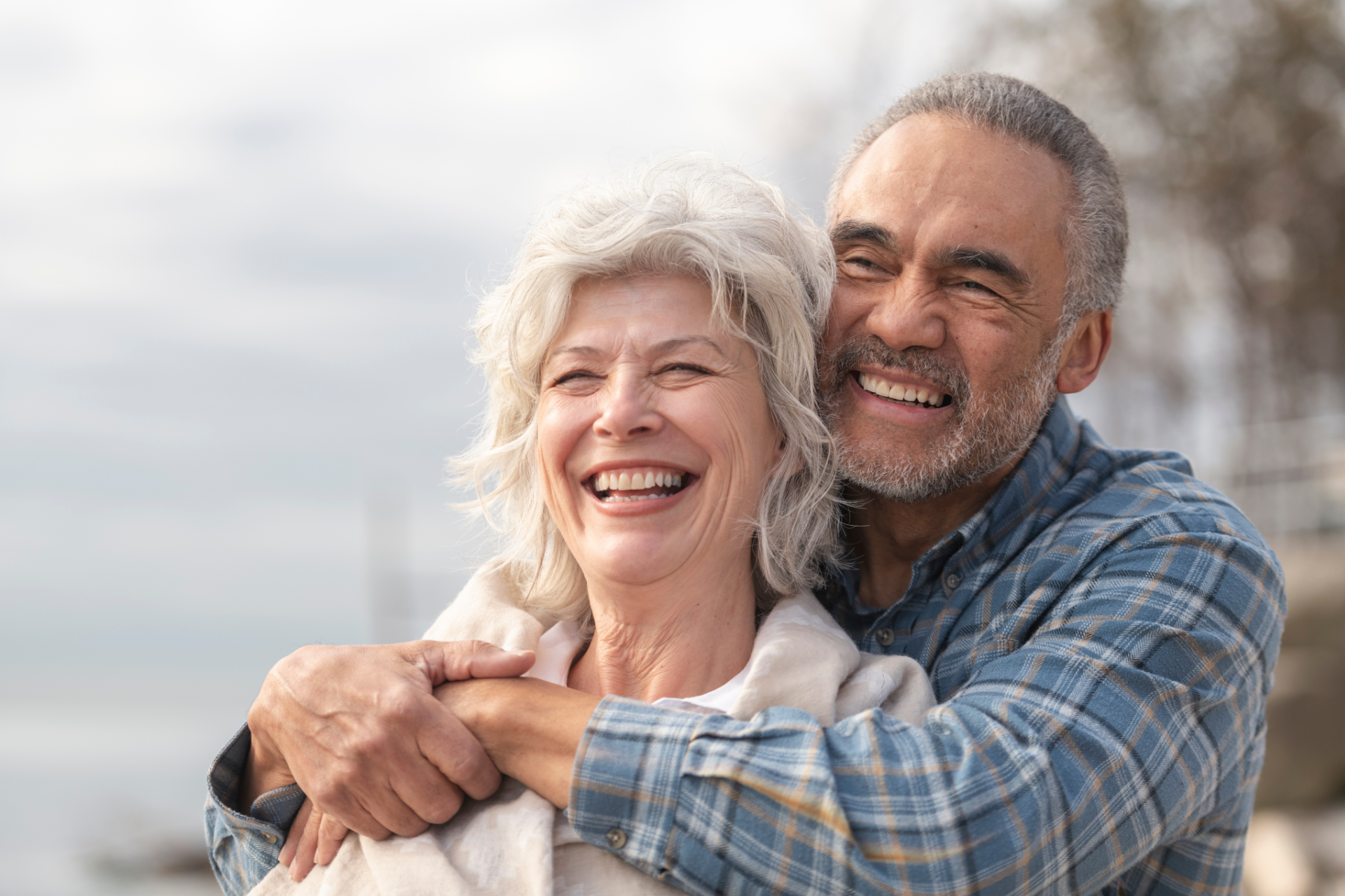 Smiling couple hugging outdoors, man behind woman. Both have gray hair.