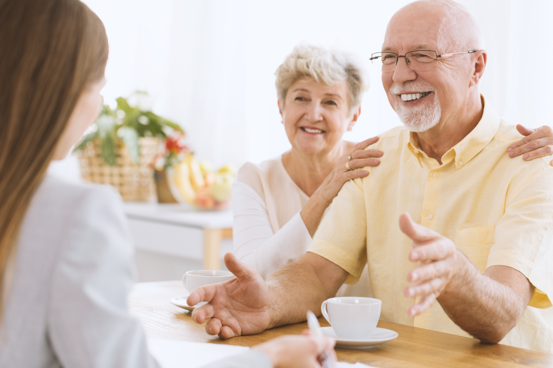 An older couple smiles, talking with a person, in a sunny room; they appear happy, and a woman is taking notes.