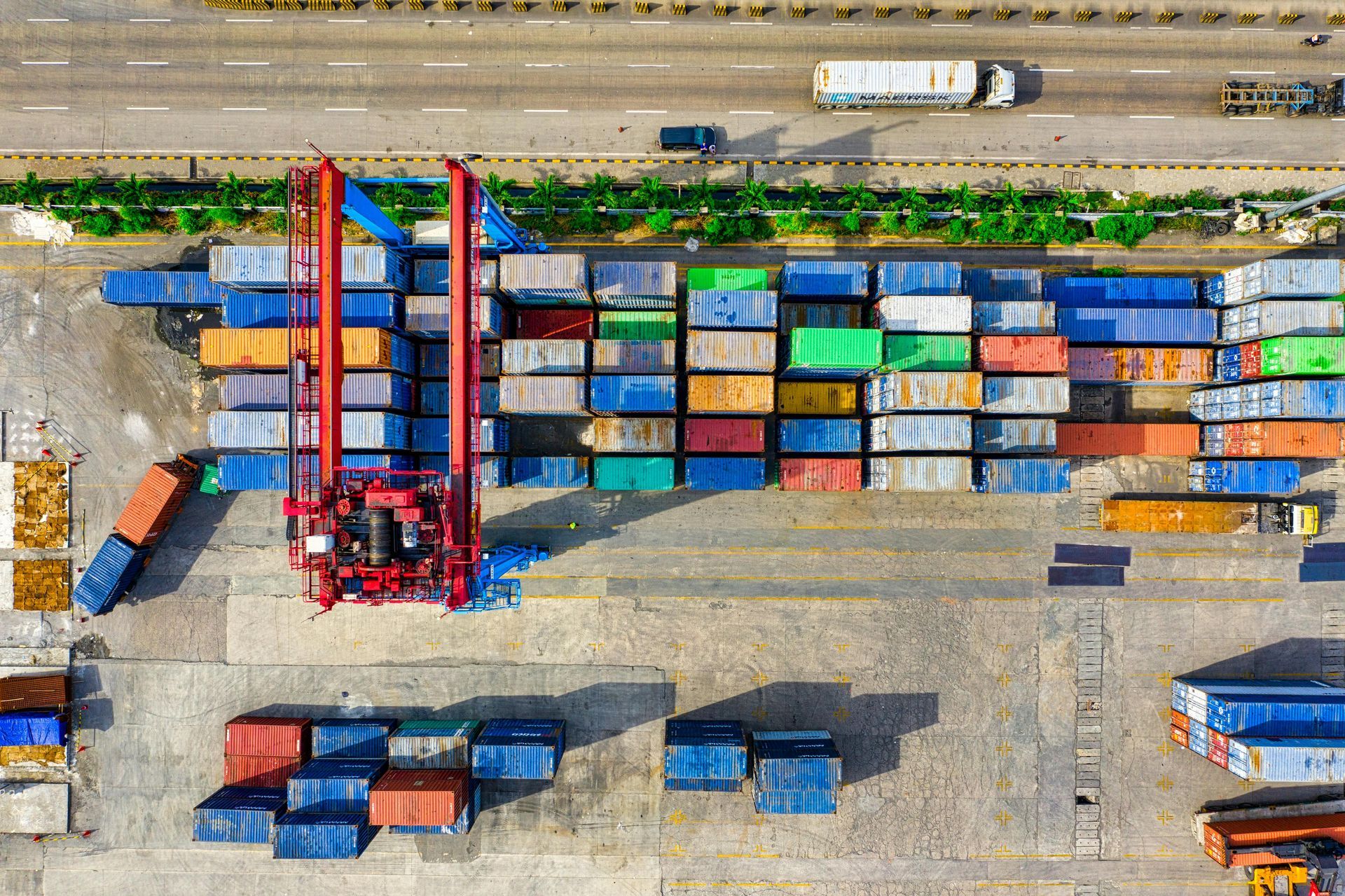 Overhead view of a cargo port with colorful shipping containers, a large crane, and trucks on a nearby highway.