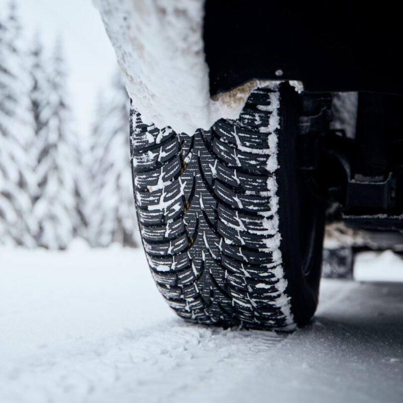 Close-up of a snow tire on a vehicle, covered in snow, set in a snowy landscape.