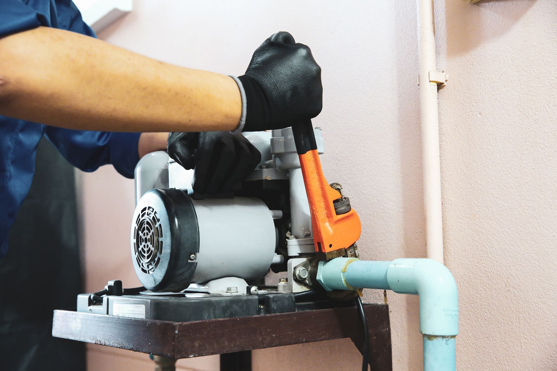 A person wearing black gloves uses an orange pipe wrench to repair a water pump and attached blue pipes.