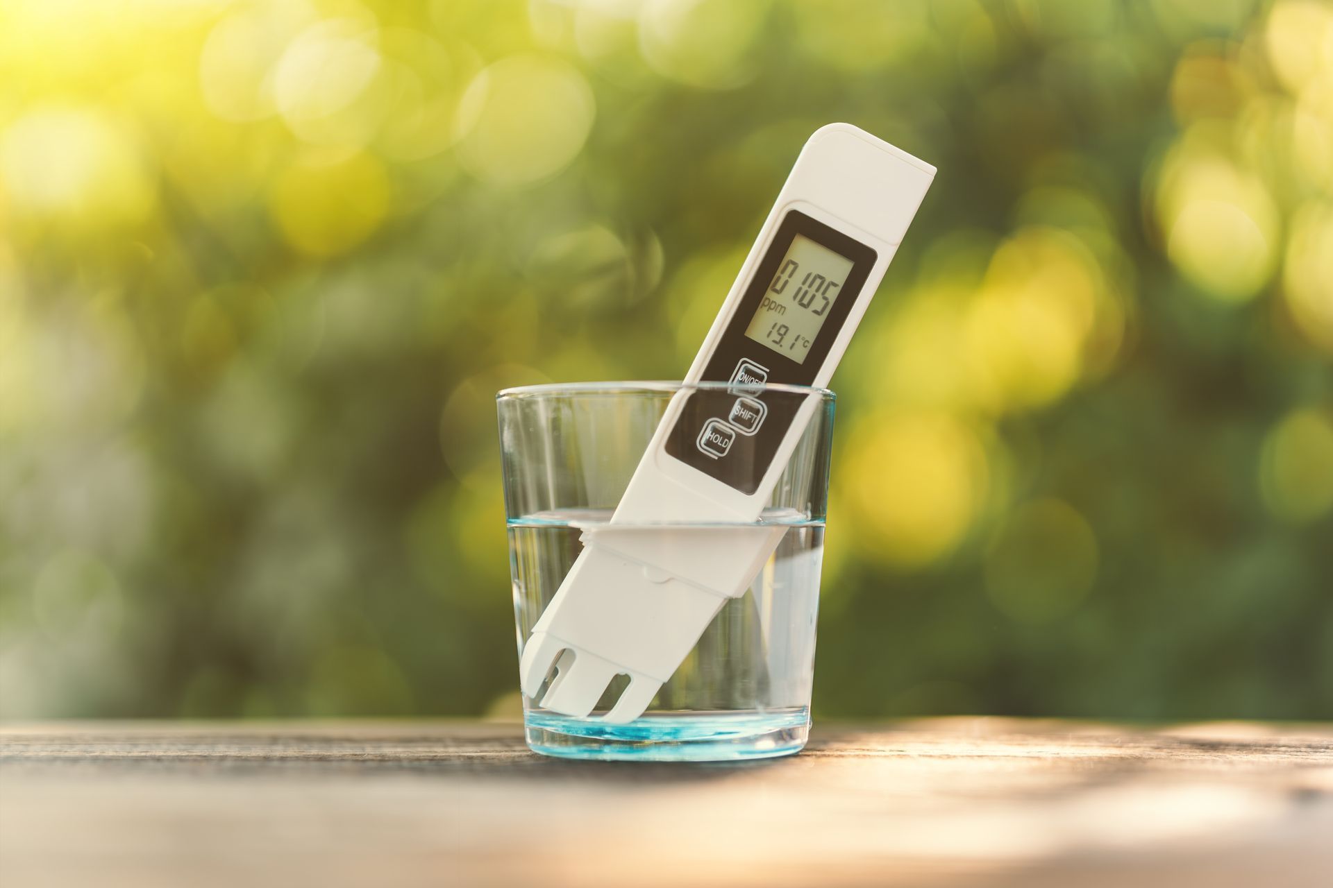 A digital water quality tester stands inside a glass of clear water against a blurred, sunlit green garden background.