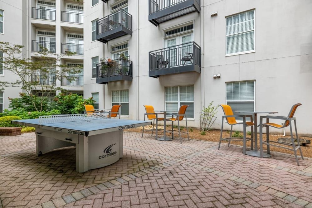 Outdoor common area of an apartment complex featuring a white Cornilleau ping-pong table, modern metal chairs with orange seats around a matching table, and landscaped plants, with multi-story apartment building facade and balconies in the background at Marq on Ponce.