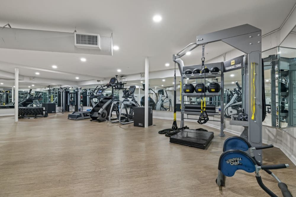 A modern, spacious gym with a variety of exercise equipment including treadmills, elliptical trainers, a rowing machine, stationary bikes, a cable machine, and multiple weight training stations. The floor is covered with wood laminate, and mirrors line the walls, enhancing the brightness of the space. The gym has a clean, organized appearance with labeled areas like 'TRAINING ZONE' at Marq on Ponce.