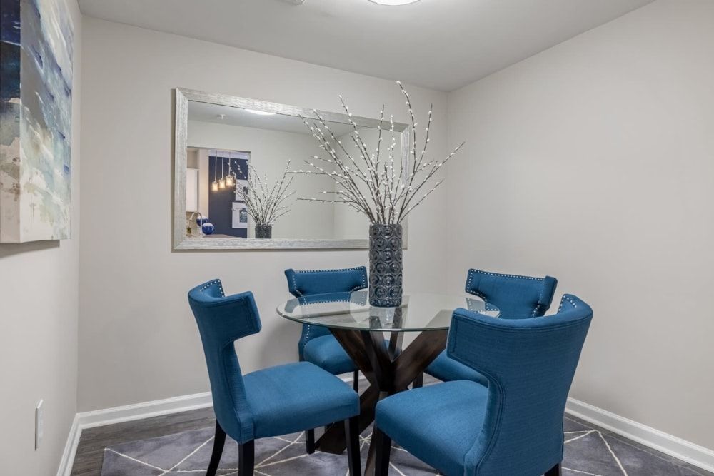 A contemporary dining room with a round glass table and four blue upholstered chairs. An abstract painting hangs on the left wall, and a large rectangular mirror on the right wall reflects part of the dining area and an adjoining room with blue decor accents. A decorative vase with white branches is centered on the table, adding a touch of elegance to the space at Marq on Ponce.