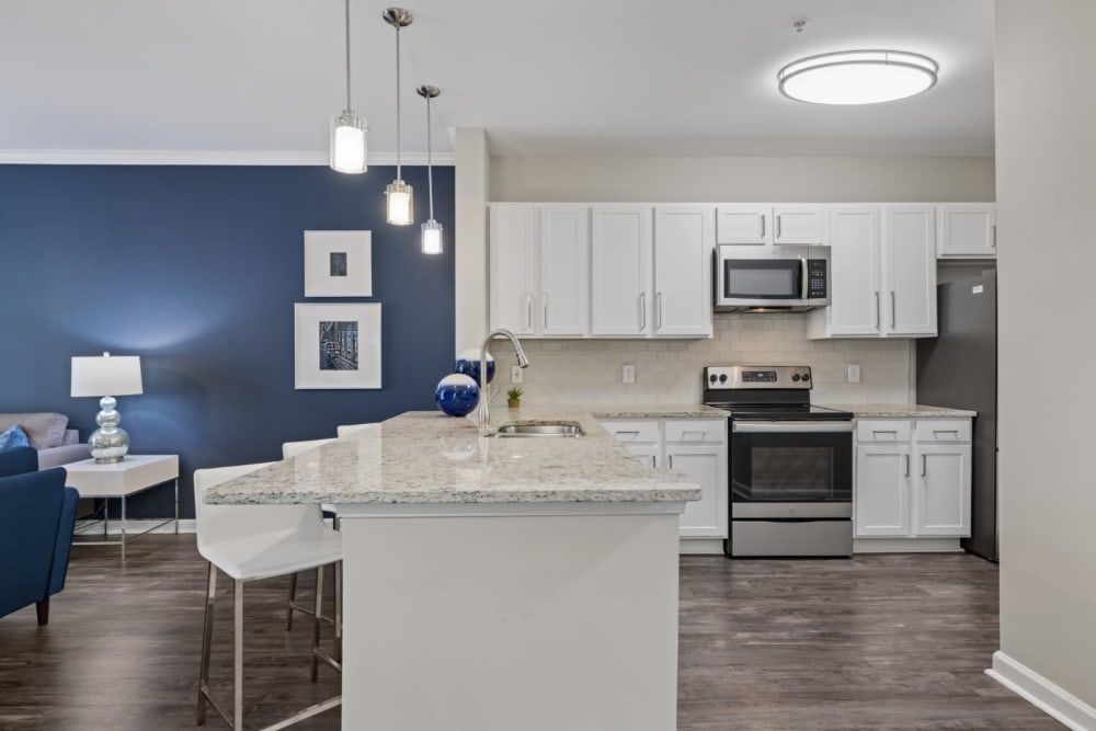 Modern kitchen with white cabinetry and stainless-steel appliances, featuring a granite countertop island. An open-plan layout reveals a living space with a blue accent wall, matching sofa, and elegant lighting fixtures. There's decorative artwork on the walls and hardwood flooring throughout at Marq on Ponce.