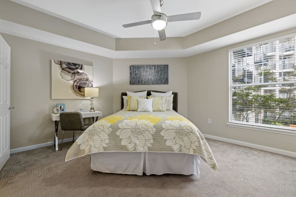 A well-lit, cozy bedroom featuring a large window with blinds, a queen-size bed draped with a yellow and white floral comforter, a gray upholstered chair at a small desk with a lamp, and decorative artwork on the walls. The room has carpet flooring, a ceiling fan, and neutral-toned walls creating a warm and inviting atmosphere at Marq on Ponce.