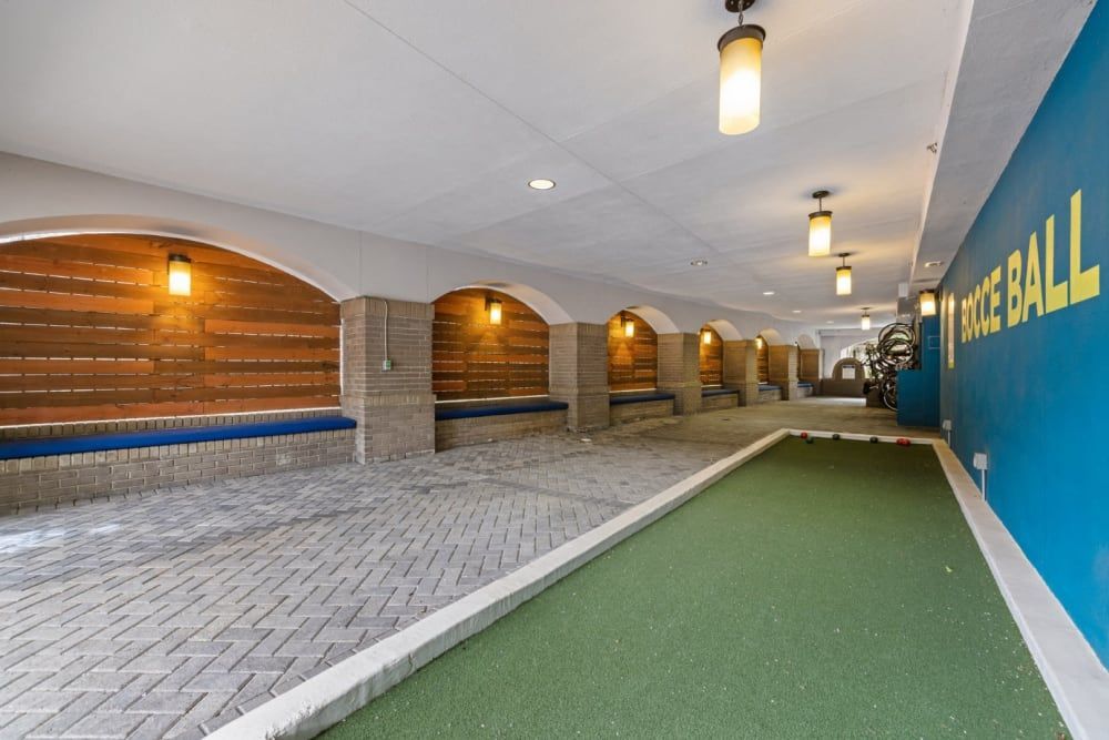 Indoor bocce ball court with a green carpeted playing area and gray brick pavement on the sides. The wall features a series of arches with recessed warm lighting, and a bold blue side wall with the text 'BOCCE BALL' in large yellow letters. A bicycle sculpture is partially visible at the end of the corridor at Marq on Ponce.