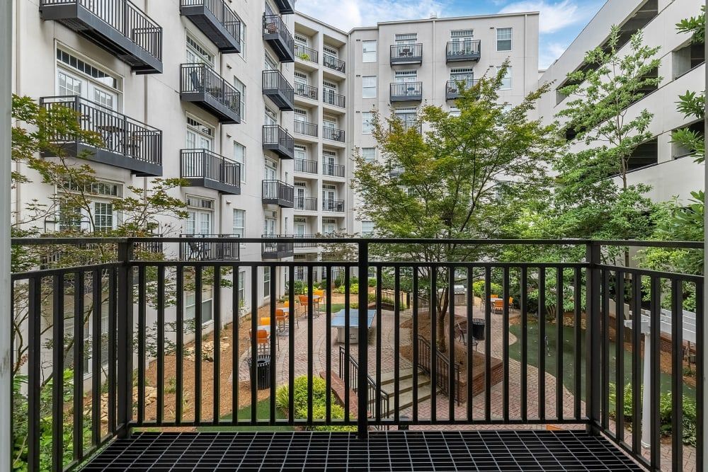 View from a balcony with black metal railings overlooking a peaceful communal outdoor area surrounded by a modern apartment complex with multiple floors, each featuring similar balconies. Green trees and landscaped vegetation add a natural touch to the urban environment at Marq on Ponce.