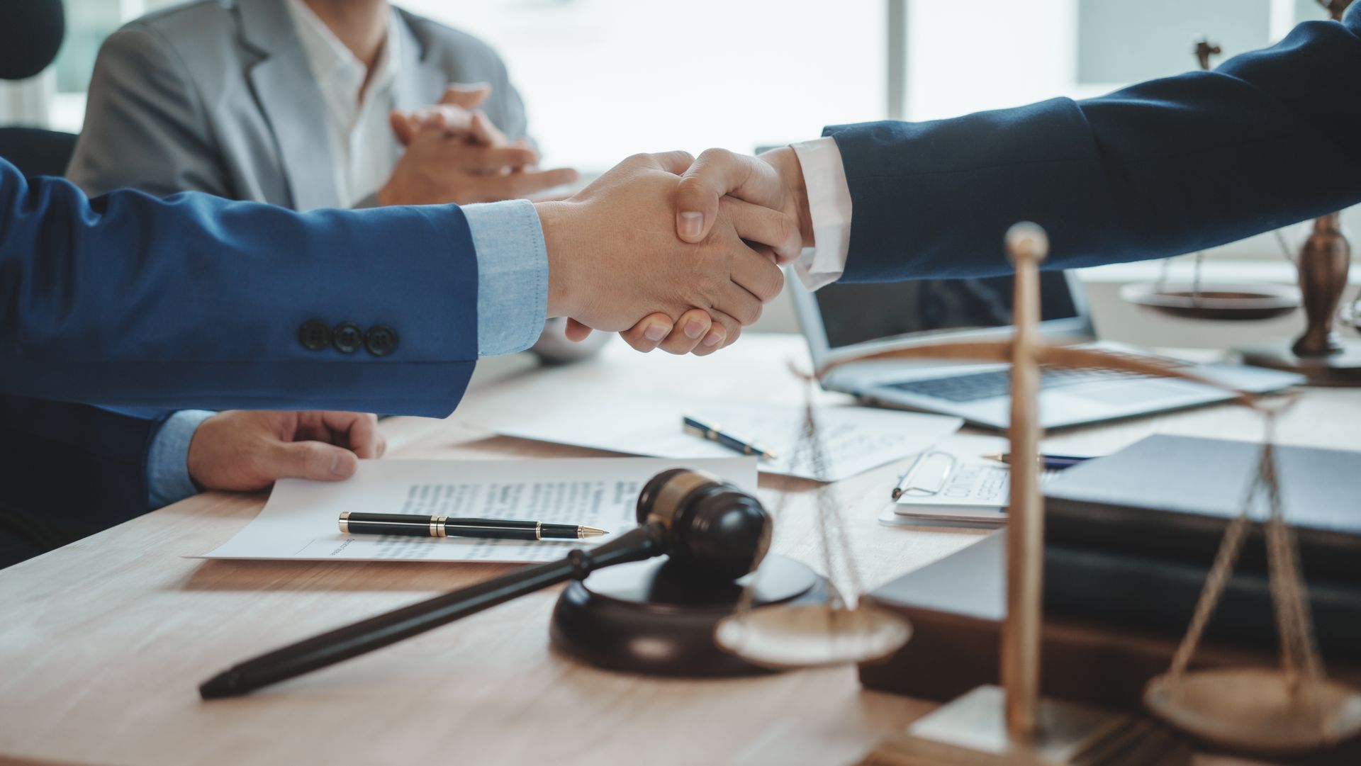 Two people shaking hands over a table with a gavel, scales of justice, and documents in a law office.