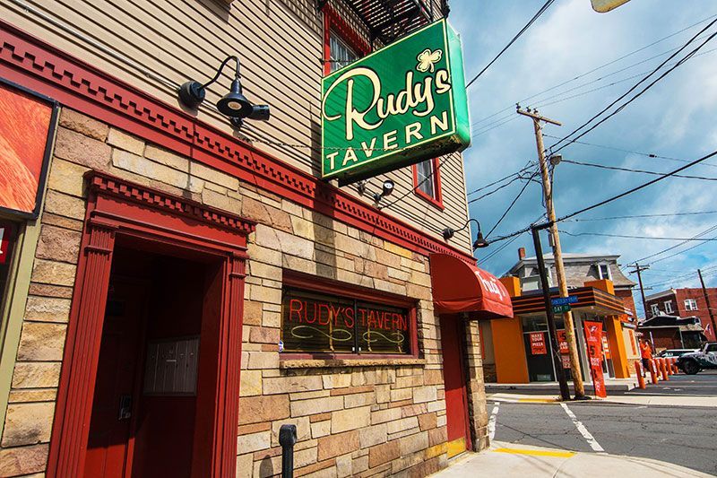 Rudy's Tavern building exterior. Tan brick facade, red trim, green sign. Street corner view with overcast sky.
