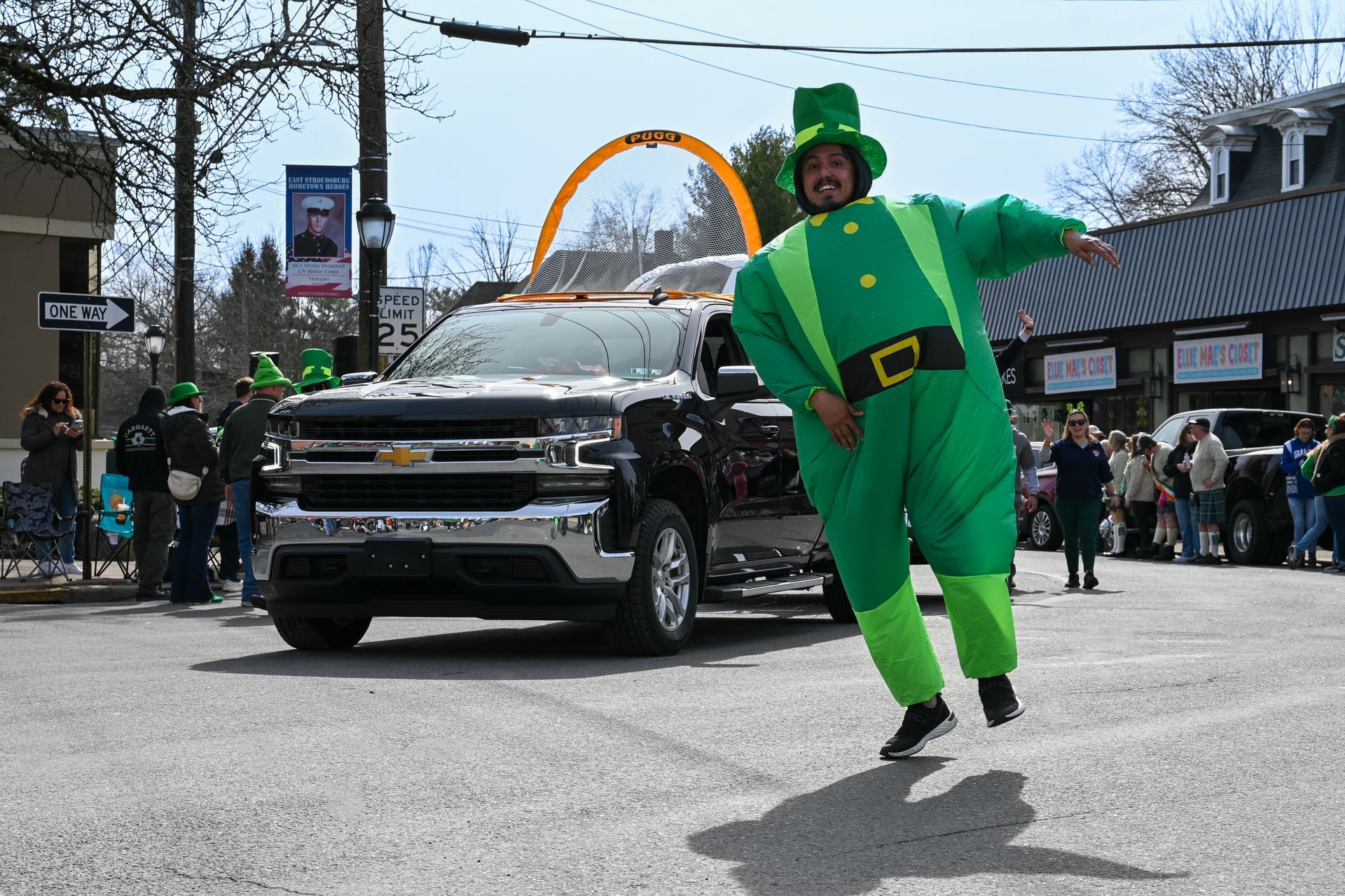Man in inflatable leprechaun costume poses beside a Chevrolet truck in a parade setting.