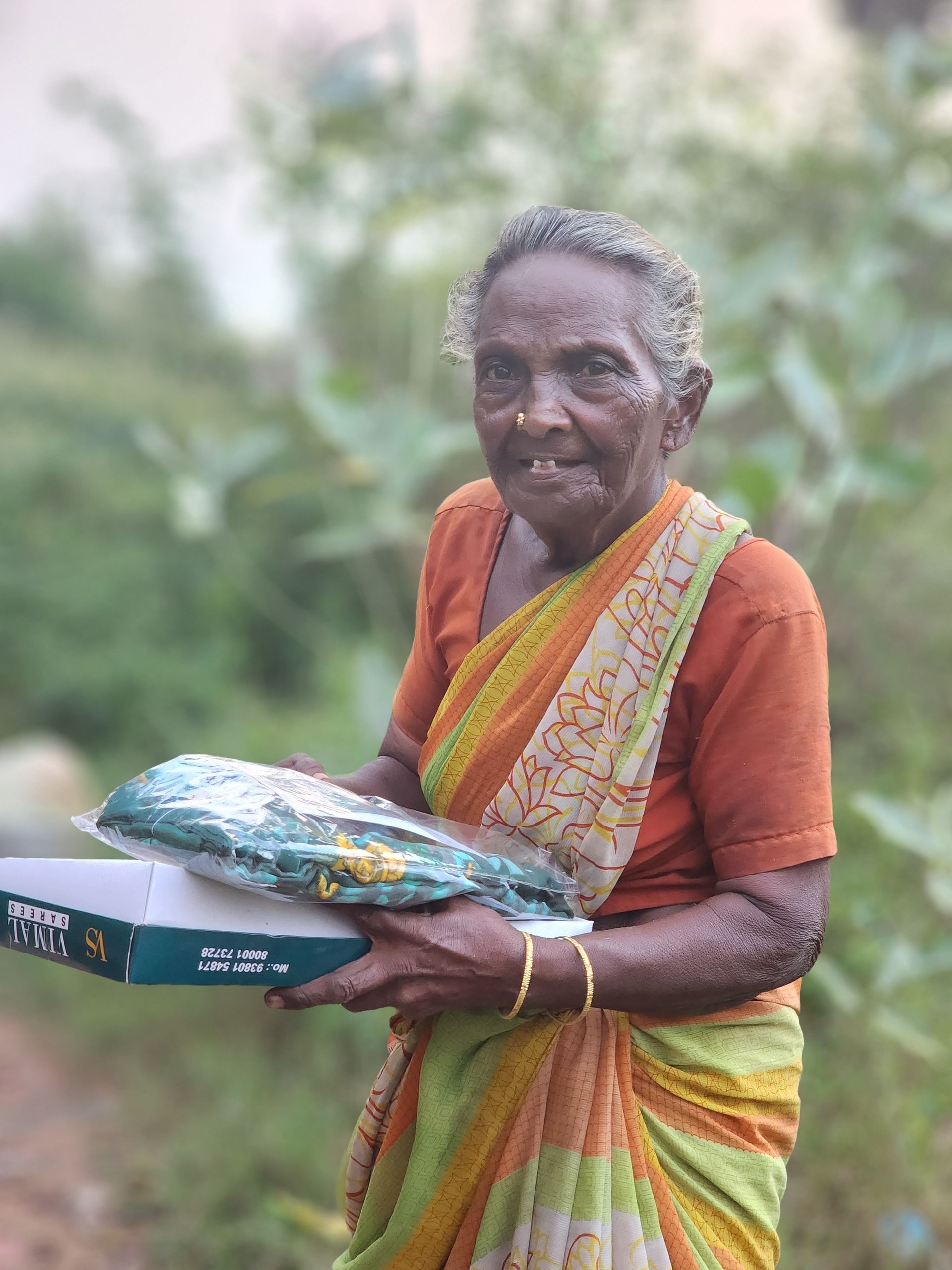 An elderly woman in a sari is holding a box and smiling.