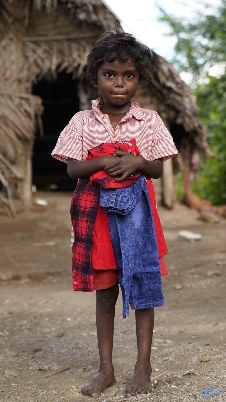 A little girl is standing in front of a hut holding a blanket.