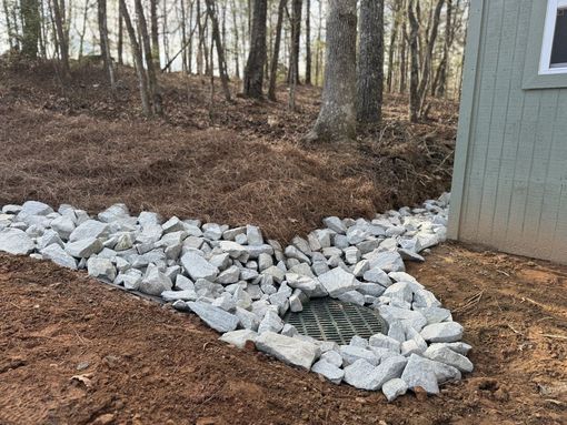 A drain is surrounded by rocks and dirt next to a shed in the woods.