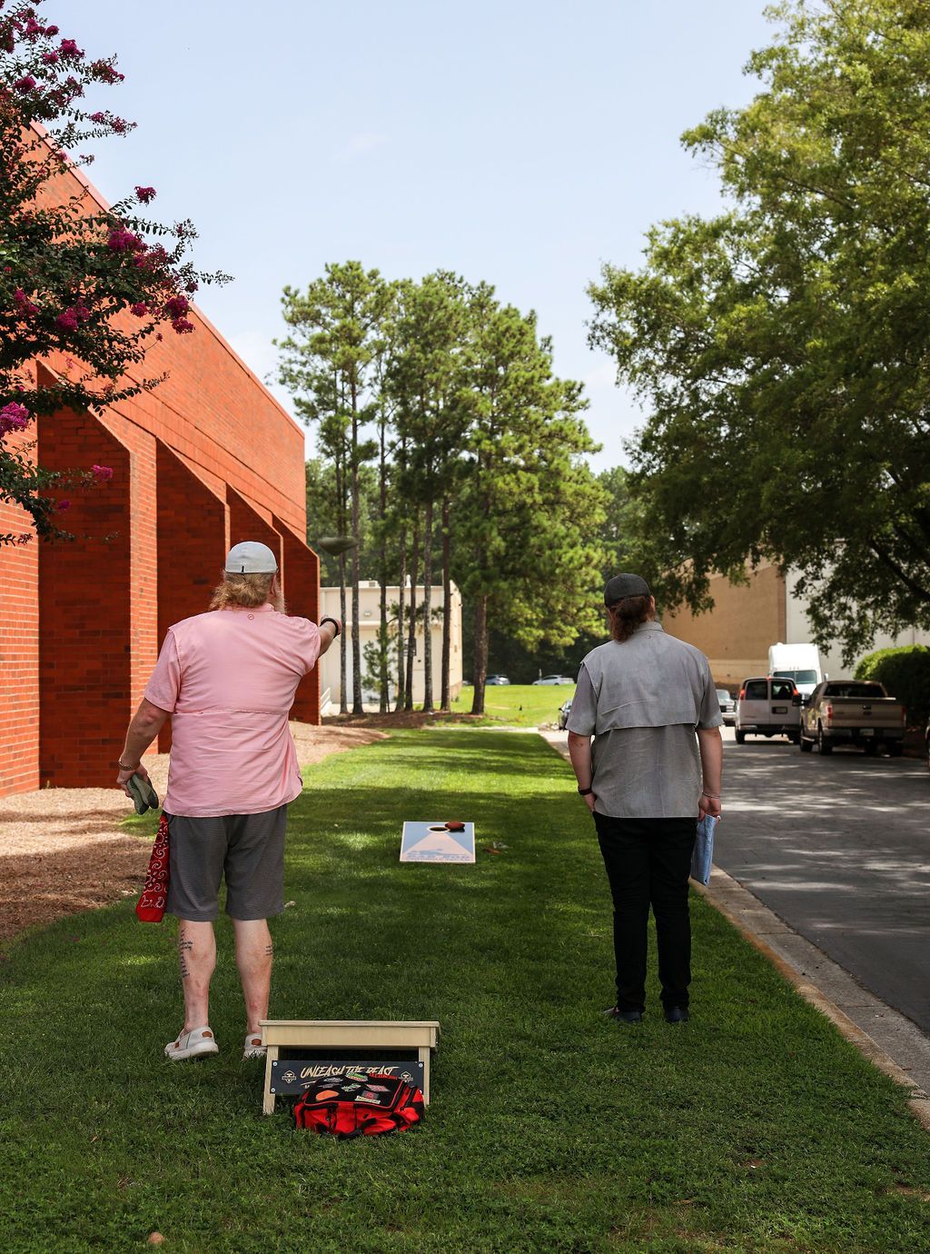 Two people are playing cornhole in the grass in front of a building.