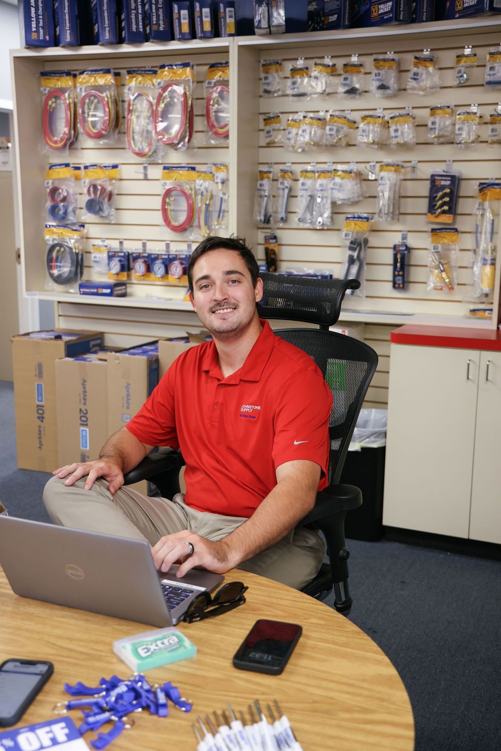 A man in a red shirt is sitting at a desk with a laptop.