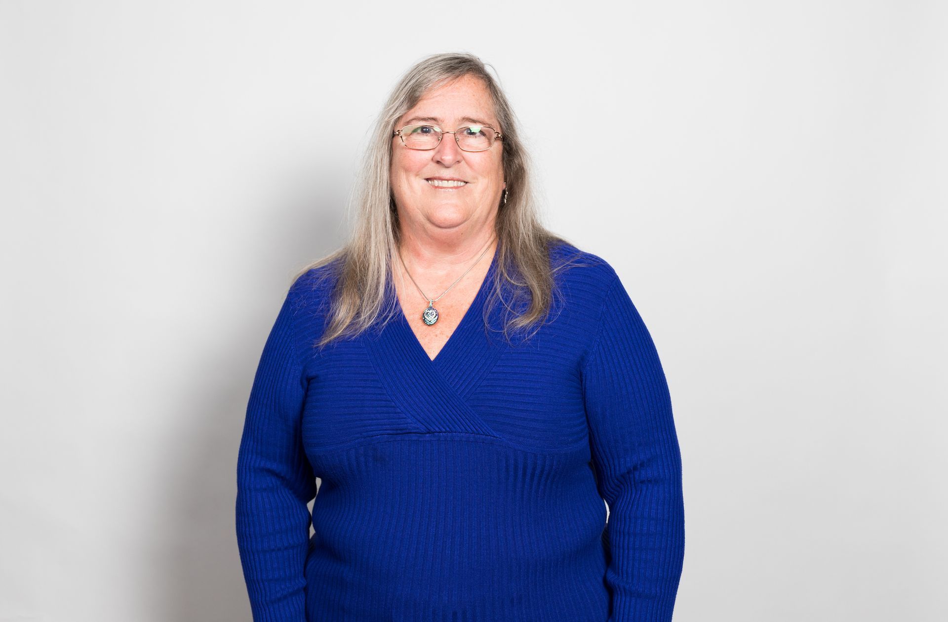 A woman wearing glasses and a blue sweater is standing in front of a white wall.