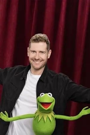 Man with Kermit the Frog puppet, arms open, smiling in front of a red curtain.