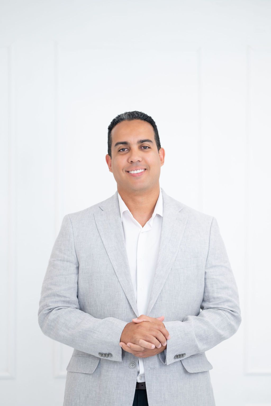 Man in light gray blazer and white shirt, smiling, standing in a white room.