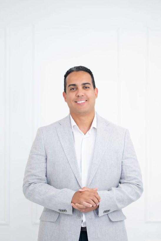 Man in light gray blazer and white shirt, smiling, standing in a white room.