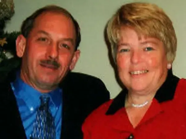 Man with mustache and woman smiling, posed together. Man in blue shirt, woman in red.