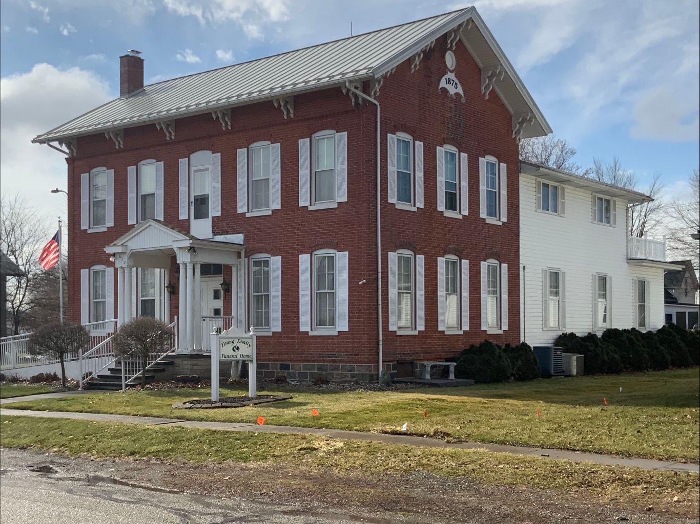 Two-story brick building with white shutters and porch, accessible ramp, and flag on the front lawn.