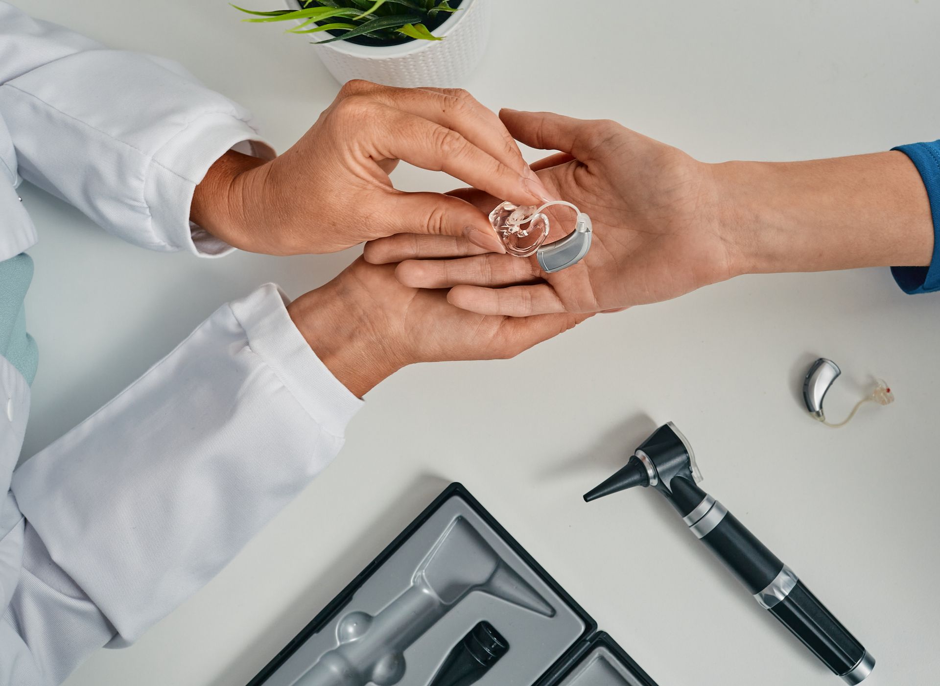 A doctor shows a hearing aid to a patient, examining it in hand. An otoscope and a box are on the white table.