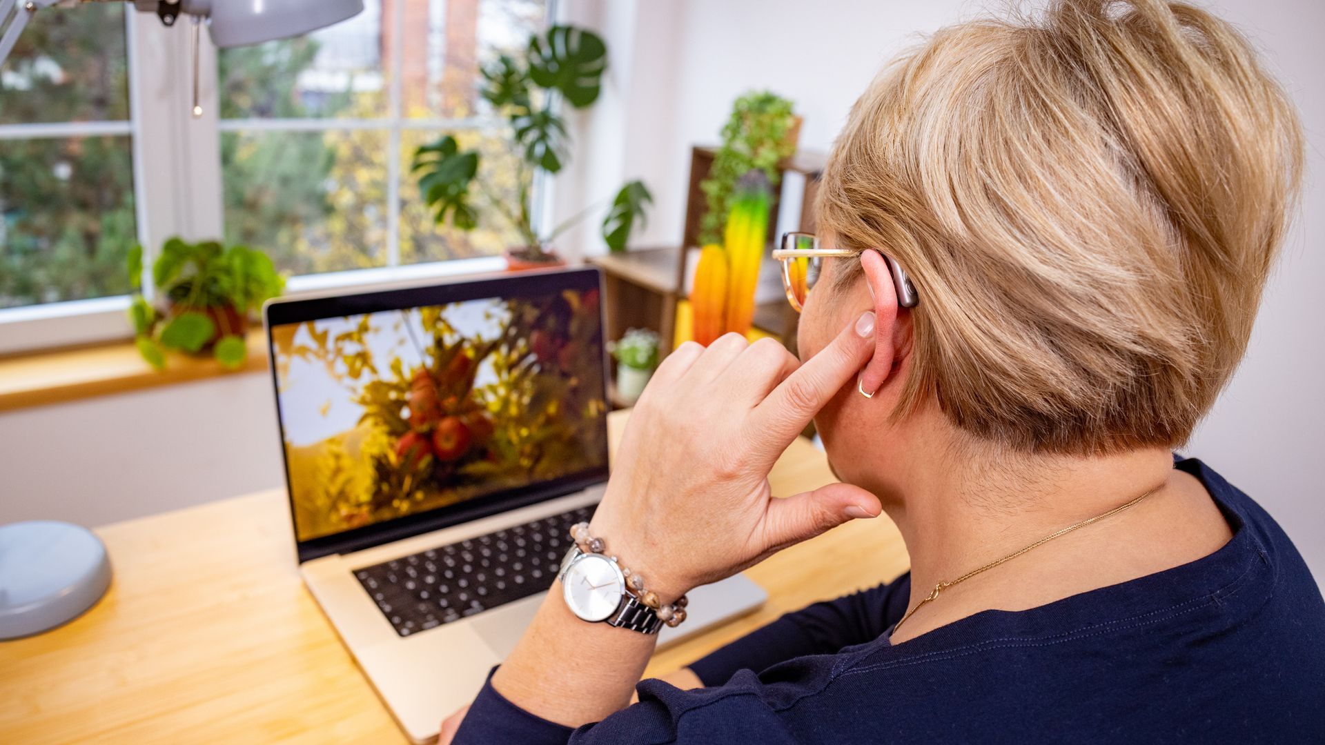 Smiling older woman with gray hair, adjusting her hearing aid against a blue background.