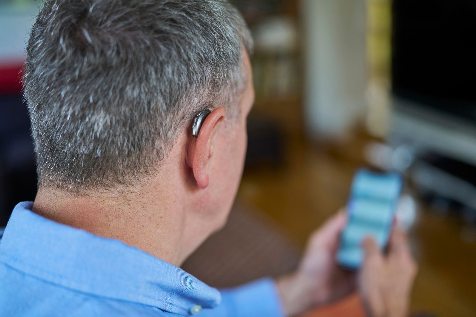 An older woman with a hearing aid and glasses uses a smartphone outdoors. She is wearing a colorful top, and her gray hair is pulled back.