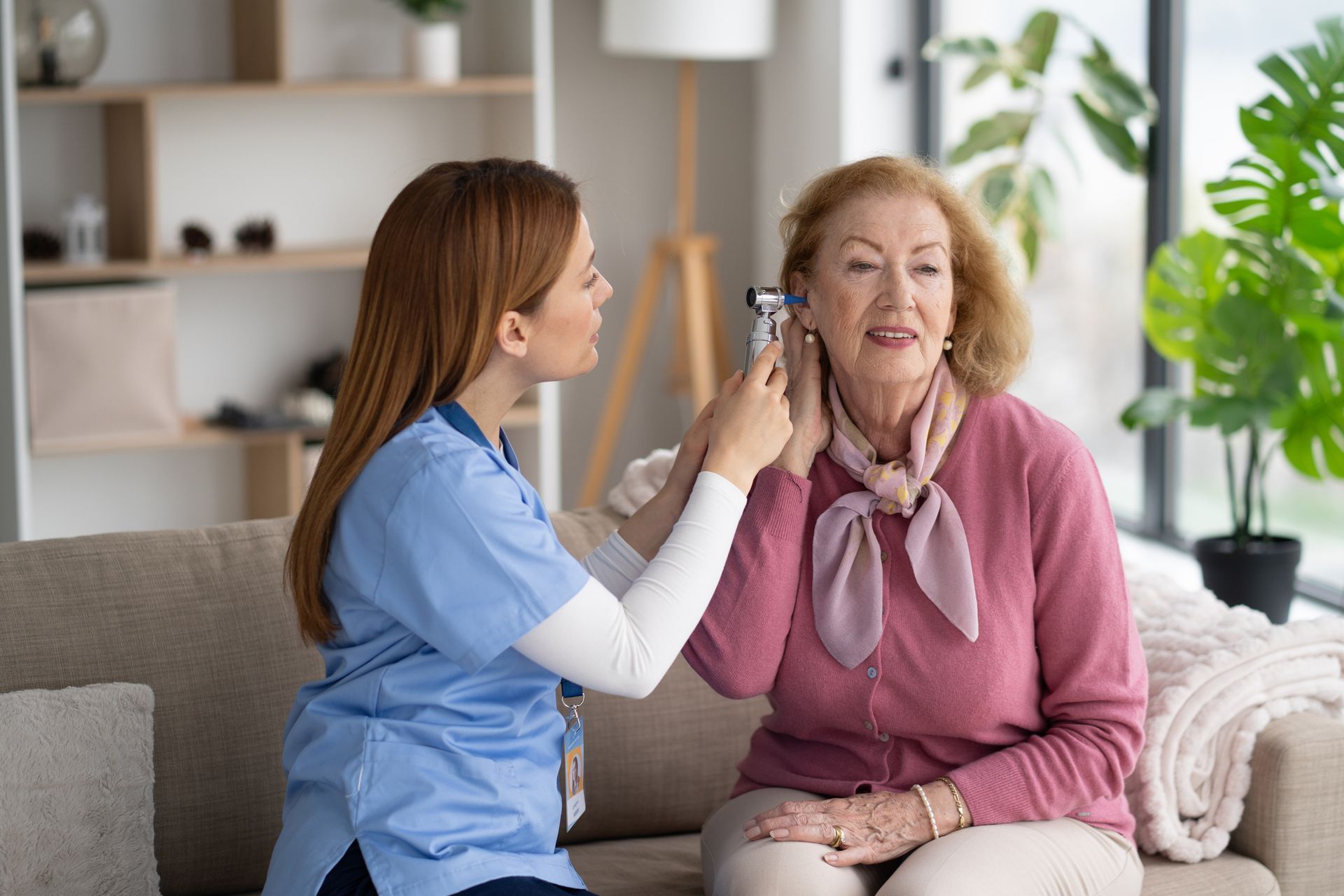 A young woman in blue scrubs examines the ear of an older woman with an otoscope while sitting on a couch in a home.