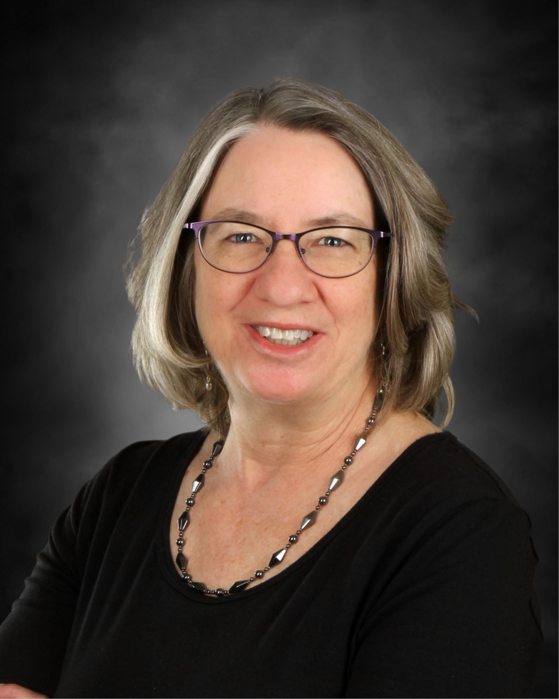 Woman with glasses and a green shirt smiles at the camera against a brown backdrop.