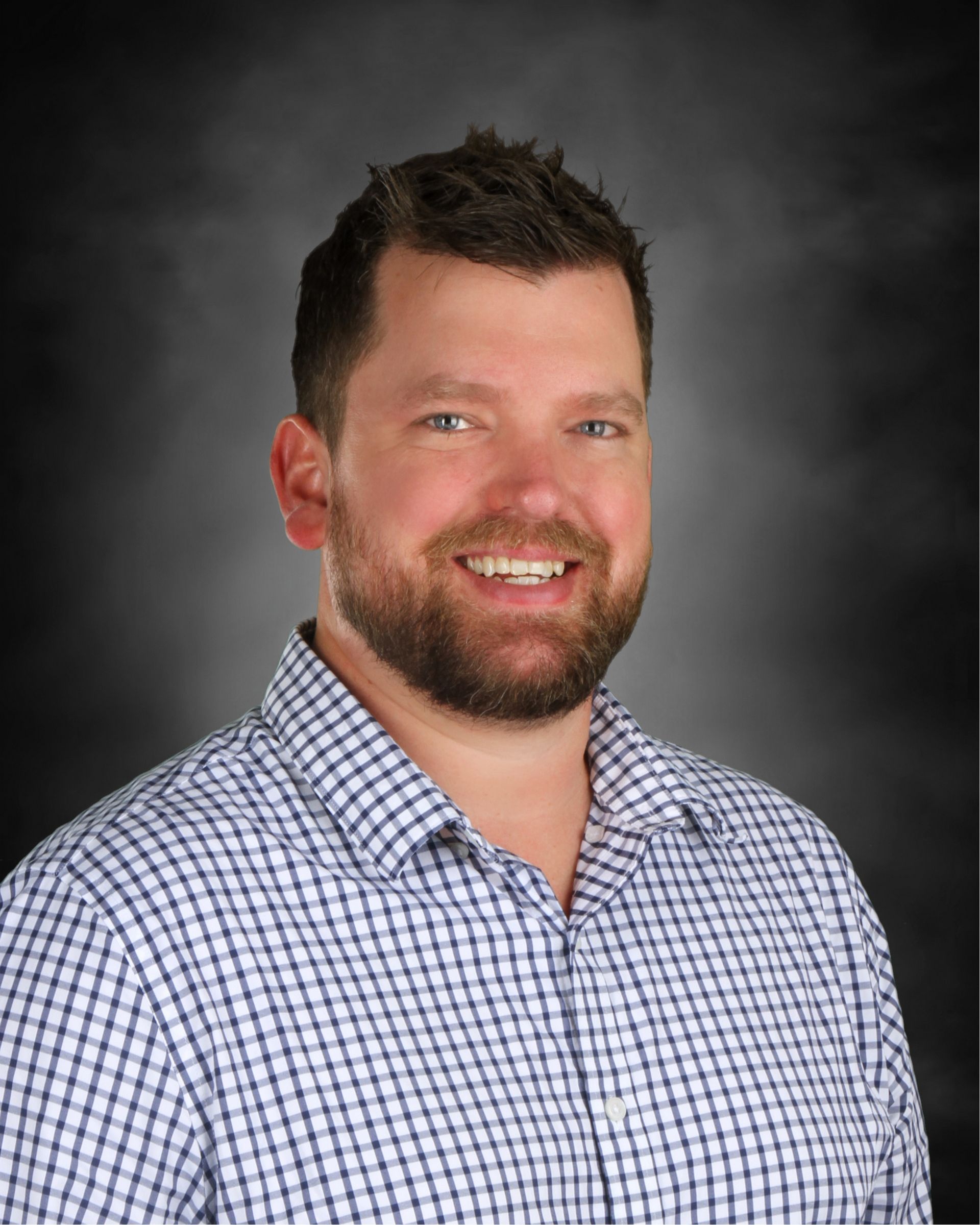 Smiling man with a beard wearing a blue and white checkered shirt against a dark background.