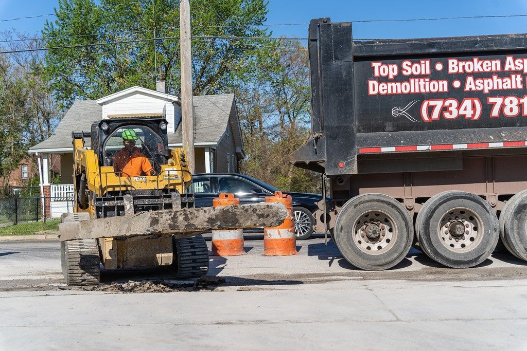 Worker on small construction vehicle beside dump truck at demolition site, with orange cones and debris