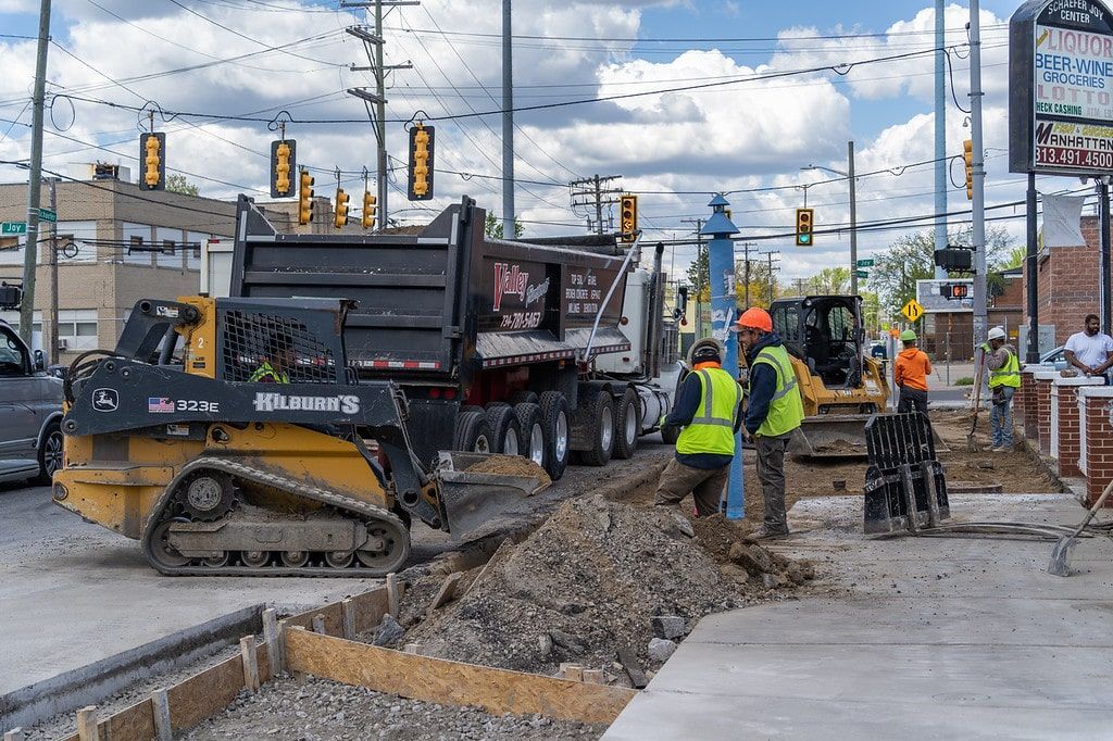 Construction crew using a skid steer and dump truck at a roadwork site with traffic lights overhead