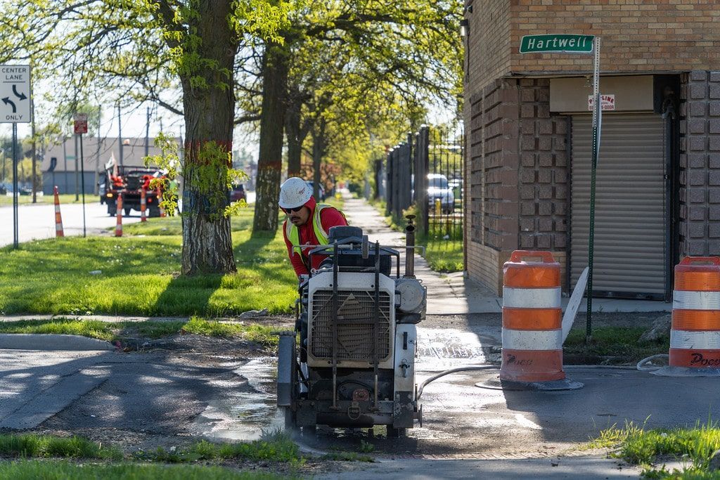 Worker using a machine on a tree-lined sidewalk near a building with orange traffic barrels