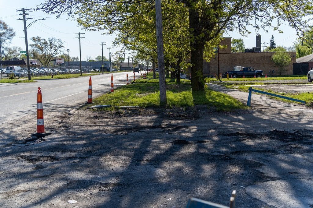 Tree-lined gravel roadside with orange traffic cones beside a paved road on a sunny day