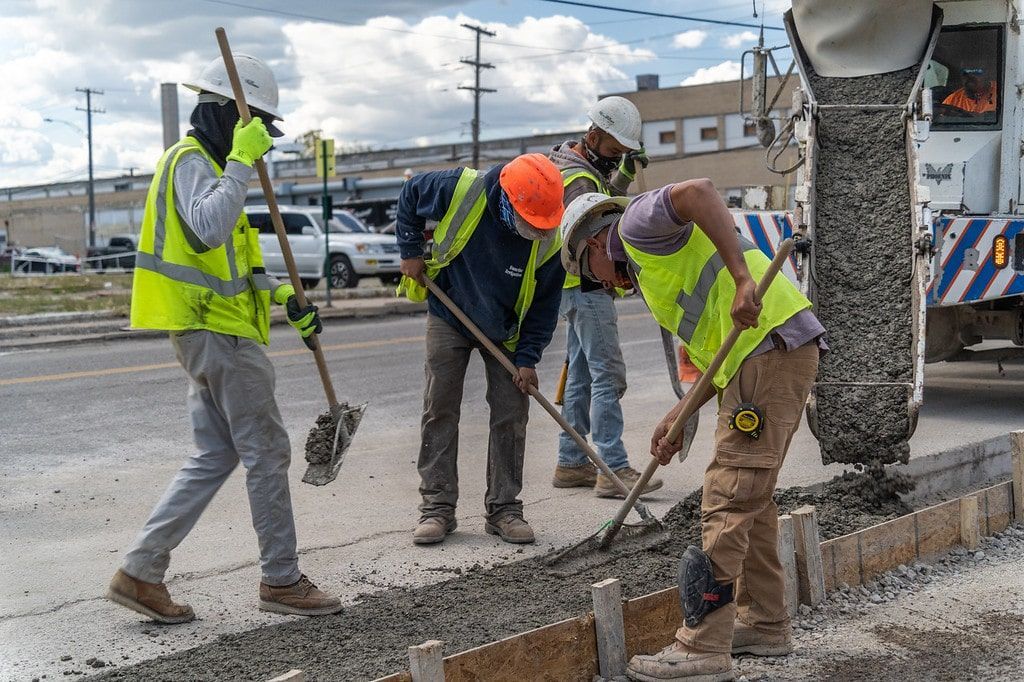 Construction workers in neon vests pour and smooth concrete on a road repair site.