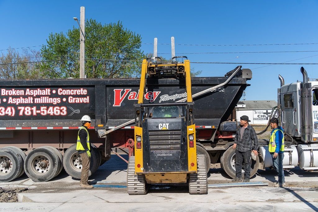 Yellow skid steer loading debris into a black dump truck at a construction site with workers nearby