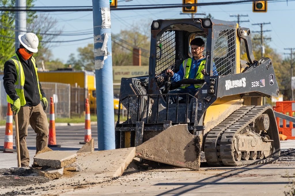 Construction workers guiding a skid-steer spreading gravel at a roadwork site with traffic cones and signals